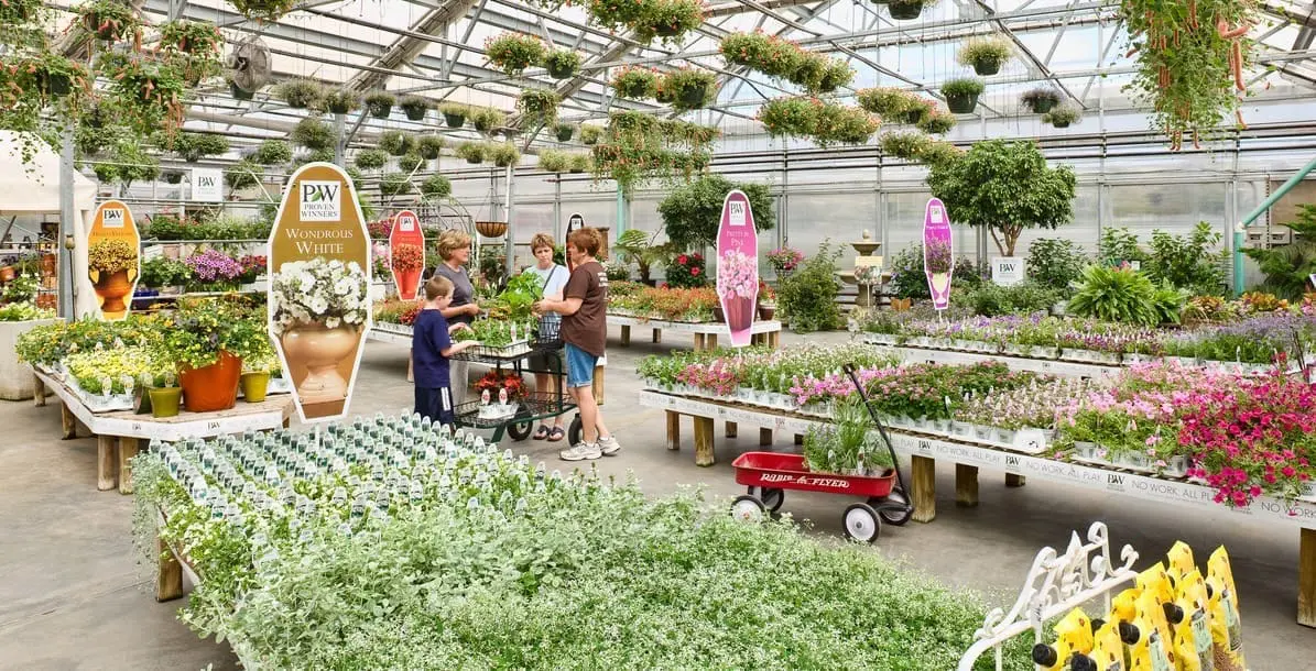 A bright retail greenhouse garden center with a family of shoppers browsing among benches full of flowering plants and a black shopping cart. Proven Winners signboards labeled 'Wondrous White' and 'Pretty in Pink' stand among the displays, with rows of hanging baskets overhead, a red Radio Flyer wagon, and a wide variety of potted plants filling the space.