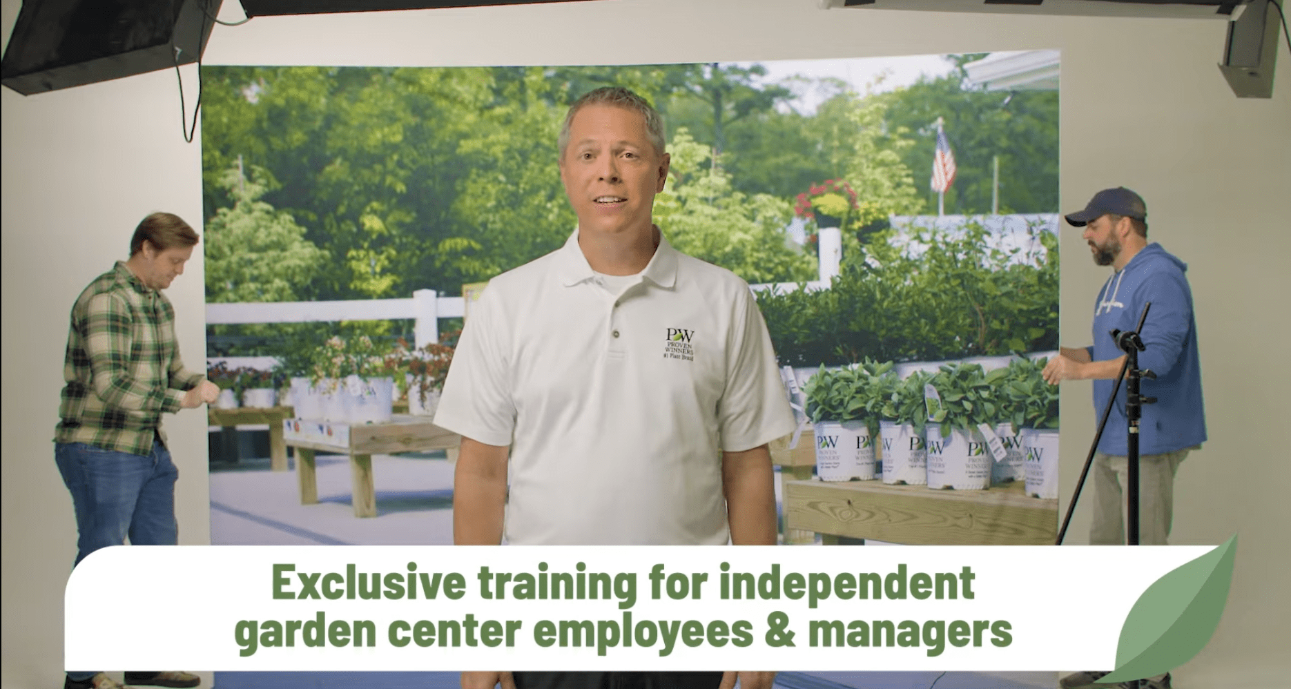 A video still from a behind-the-scenes shoot showing a man in a white Proven Winners polo shirt speaking to camera in front of a large printed backdrop of a garden center. Two crew members adjust the backdrop and equipment on either side, with studio lighting overhead. Green text across the bottom reads 'Exclusive training for independent garden center employees & managers.